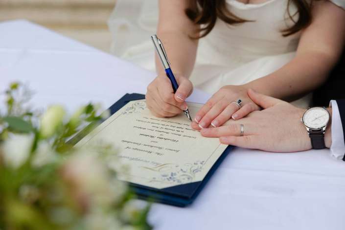 A bride signs a marriage certificate with a pen while her partner’s hand rests nearby, both wearing wedding rings.