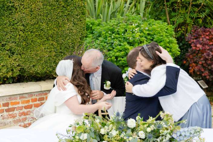 A wedding party shares embraces in an outdoor garden scene, with a bride in white and floral arrangements in the foreground.