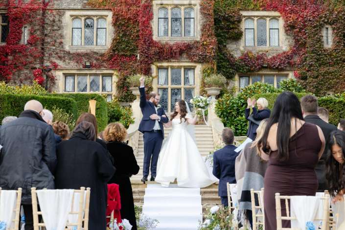 Bride and groom celebrate on the ivy-covered stone steps of a historic building as guests applaud.