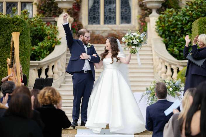 Bride and groom celebrate on an elegant outdoor staircase, smiling and raising their arms as guests applaud.