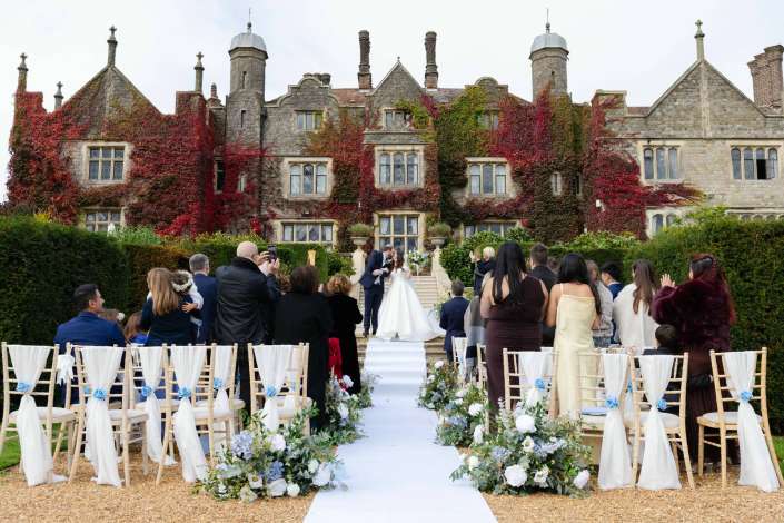 Bride and groom kiss at an outdoor wedding ceremony in front of an ivy-covered stone mansion, with guests seated along a white aisle.