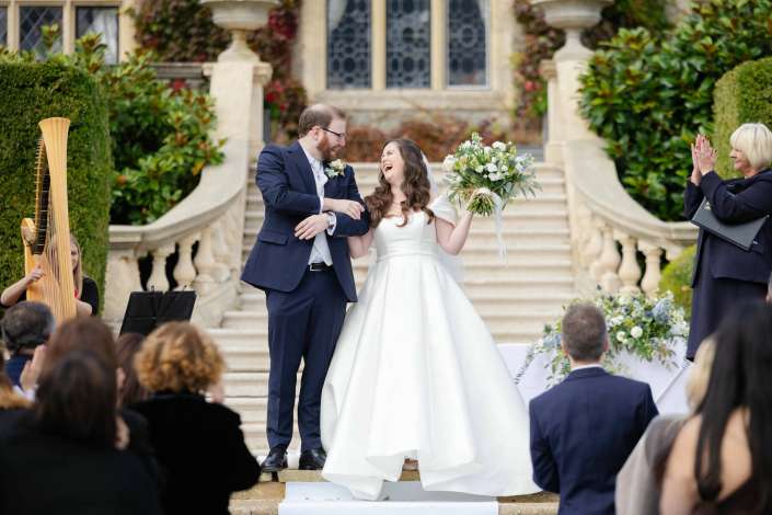 Bride in a white gown and groom in a navy suit share a joyful moment on a stone staircase during an outdoor wedding as guests applaud.