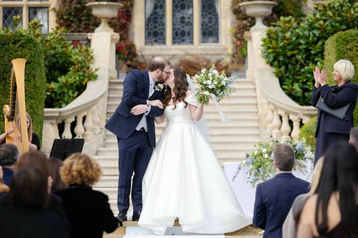 Bride and groom kiss on the stone staircase of an outdoor wedding as guests applaud.