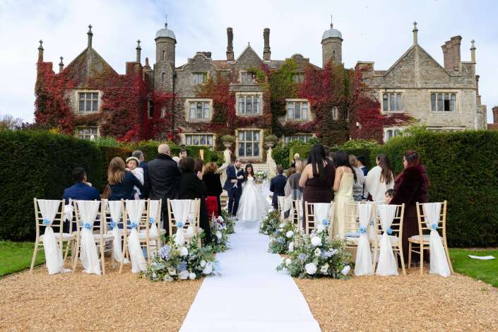 Outdoor wedding ceremony in front of an ivy-covered stone manor, with a white aisle runner, floral arrangements along the aisle, and guests gathered.