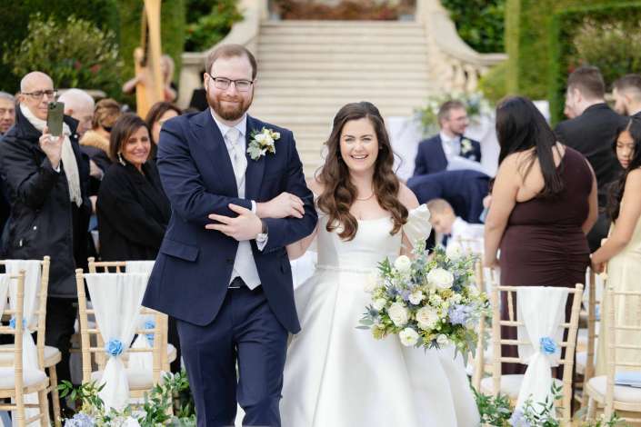 A smiling bride in a white gown holding a bouquet walks arm in arm with a groom in a navy suit down an outdoor wedding aisle, with guests and floral decor in the background.