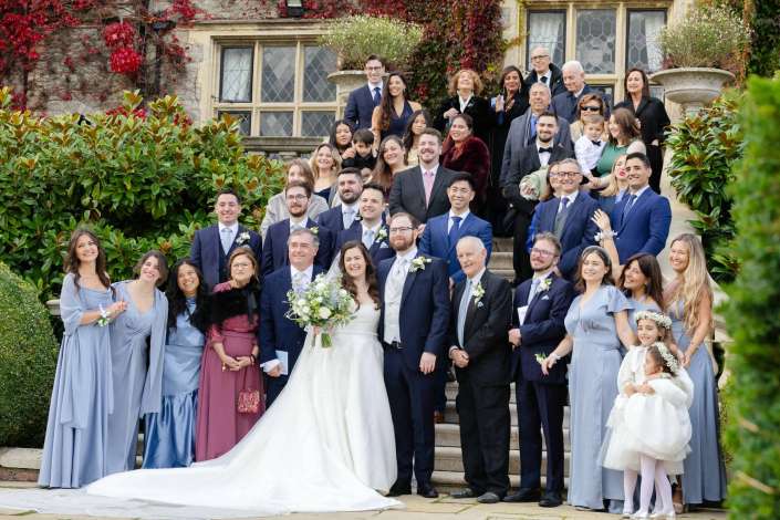 The bride and groom stand at the center of a large wedding party on a stone staircase outside an ivy-covered historic building.