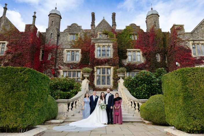 A wedding party stands on a grand stone staircase in front of an ivy-covered, castle-like mansion.