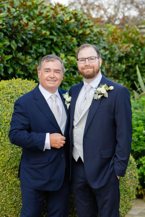 Two men in navy suits with boutonnieres pose together outdoors at a wedding.