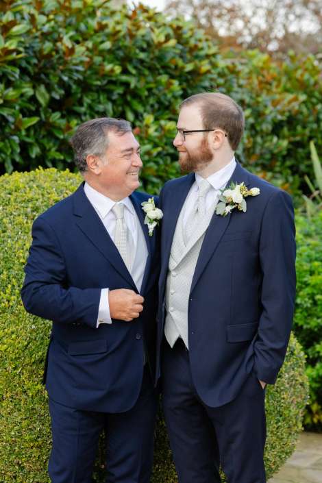 Two men in navy suits with light ties and boutonnieres stand in a garden, smiling at each other.