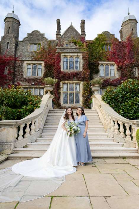 Bride in a white wedding gown holding a bouquet poses with a bridesmaid in a light blue dress on stone steps in front of an ivy-covered castle-like mansion.