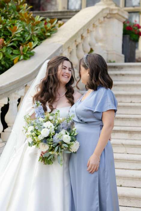 Bride in a white wedding gown with veil holding a white and blue bouquet stands with a bridesmaid in a pale blue dress on a stone staircase.