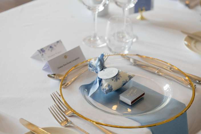 Close-up of an elegant place setting with a gold-rimmed charger, blue patterned napkin tied with a ribbon, and a name card on a white tablecloth.