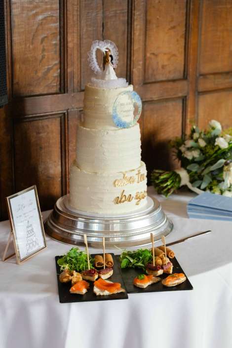 A tall white multi-tier wedding cake with a decorative topper on a silver pedestal, accompanied by an assortment of hors d'oeuvres on a slate platter.