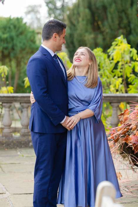 A couple dressed in blue stands in a garden, smiling at each other while holding hands.