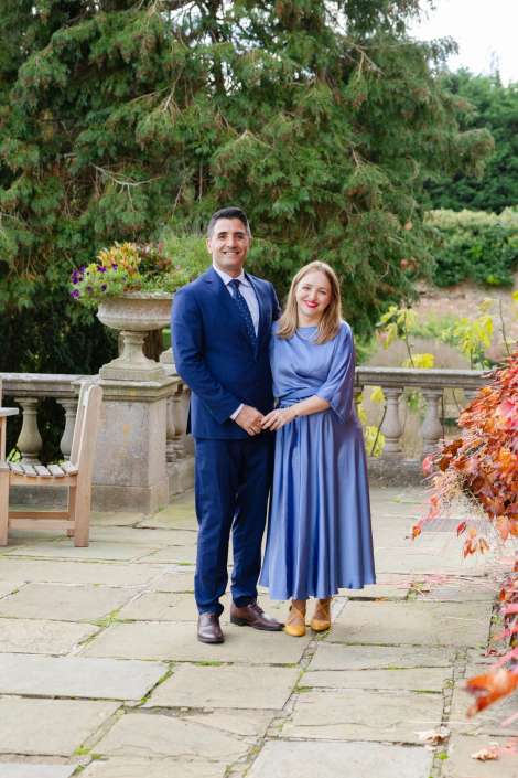 Smiling couple stands on a stone patio, the man in a blue suit and the woman in a light blue dress, with lush greenery and a stone balustrade behind them.