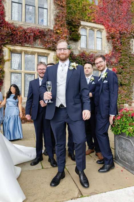 Groom and groomsmen in navy suits pose outside an ivy-covered stone building, with a bridesmaid in a blue dress nearby.