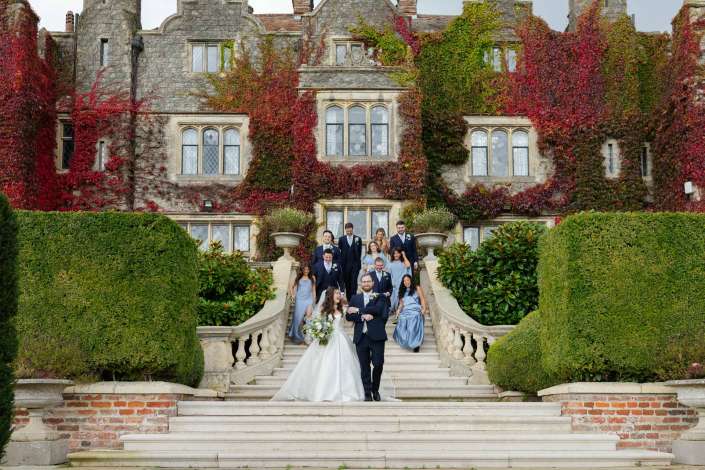 Bride and groom with their wedding party descend a grand stone staircase in front of an ivy-covered historic mansion.