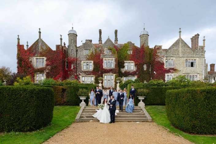A wedding party in formal attire descends the stone steps of an ivy-covered historic manor.
