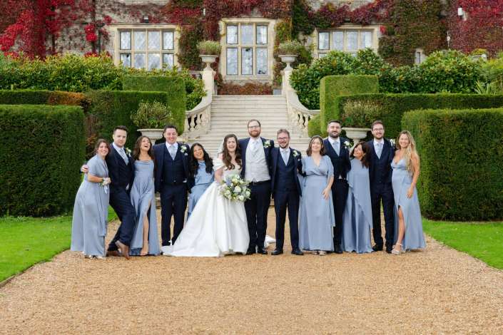 Bride and groom stand with their wedding party on a gravel path in front of an ivy-covered mansion with a grand stairway.