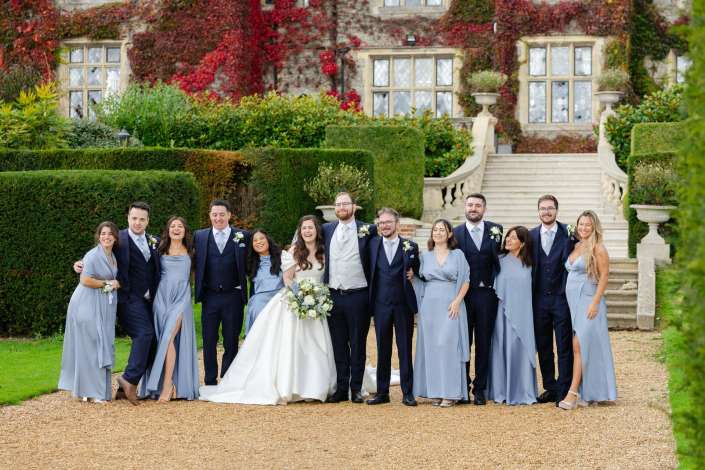 Bridal party posing outdoors in front of an ivy-covered stone mansion, with the bride and groom flanked by bridesmaids in pale blue dresses and groomsmen in dark suits.