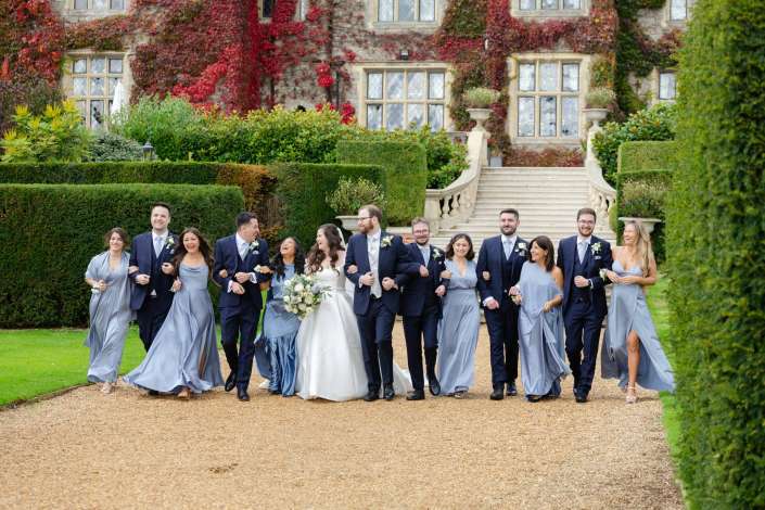 Bride and groom walk arm-in-arm with their wedding party in light blue gowns and navy suits along a gravel path in front of an ivy-covered stone mansion.