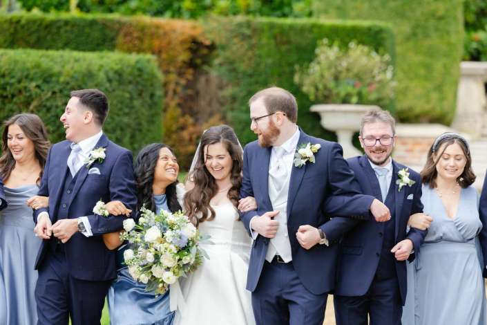 Bride in white and groom in a navy suit walk arm in arm with bridesmaids in light blue dresses and groomsmen in navy suits in a lush garden.