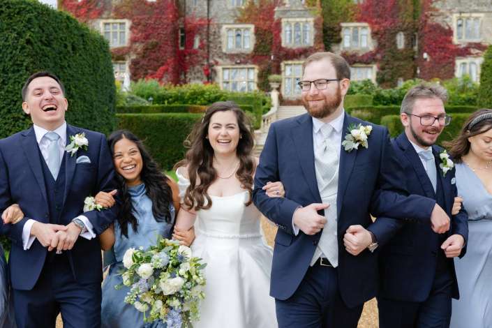 Joyful wedding party walking arm in arm outdoors, with the bride in a white gown flanked by bridesmaids in blue and groomsmen in navy suits, in front of an ivy-covered historic mansion.