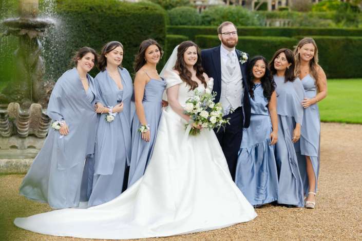 Bride in a white gown holding a bouquet poses with the groom in a suit and bridesmaids in light blue dresses outdoors by a fountain.