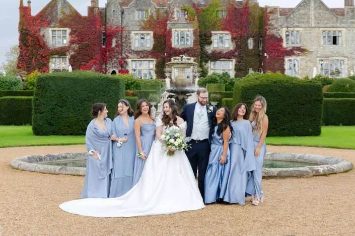 Bride and groom pose with bridesmaids in light blue dresses beside a fountain in a formal garden in front of an ivy-covered stone manor.