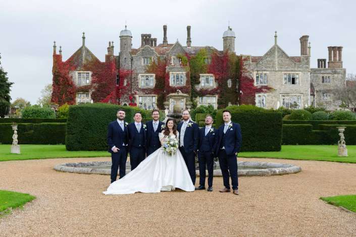 Bride in a white wedding gown stands with six groomsmen in navy suits in front of an ivy-covered historic stone manor with a circular fountain and trimmed hedges.