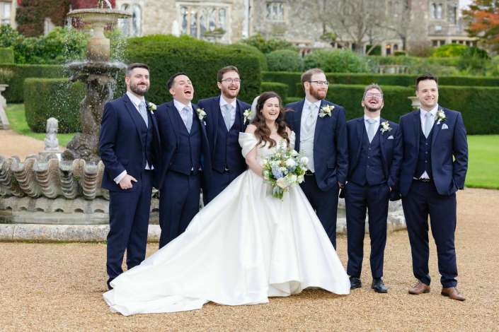 Bride in a white gown holding a bouquet stands with six groomsmen in navy suits, laughing in a formal garden with a fountain.