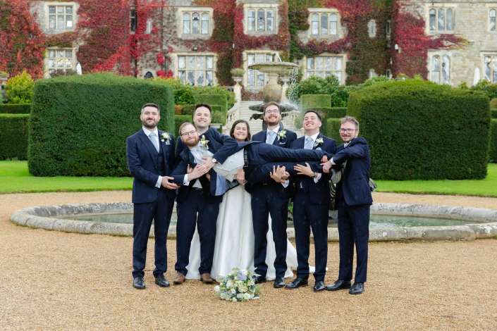 Bride in a white wedding dress is carried horizontally by her groomsmen in navy suits in front of a fountain, with an ivy-covered mansion and neatly trimmed hedges in the background.