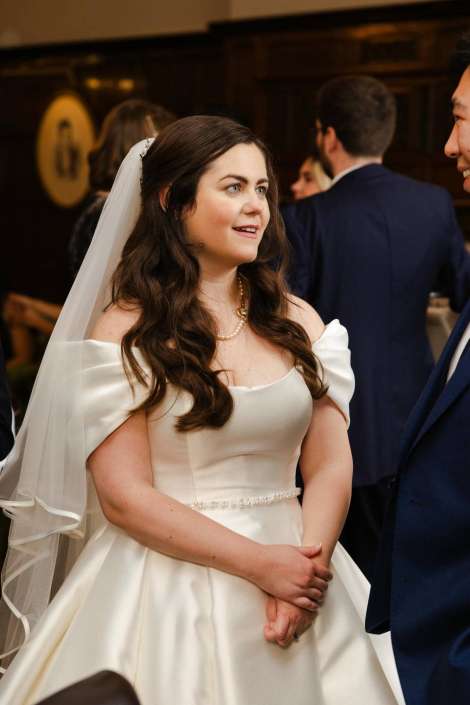 Bride in an off-the-shoulder white wedding gown with a veil and pearl necklace, standing with her hands clasped.