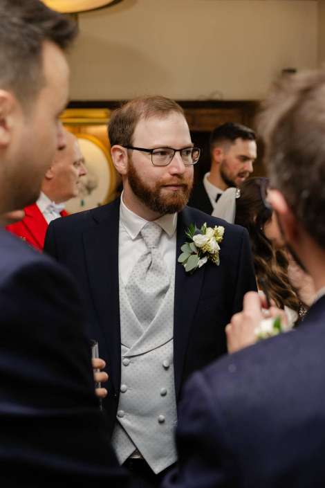 Bearded man wearing glasses in a navy suit with a silver waistcoat and boutonniere at a formal event.