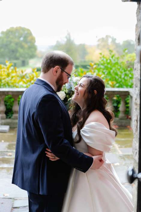 A bride in a white off-the-shoulder wedding gown embraces a groom in a dark suit, smiling at each other outdoors.