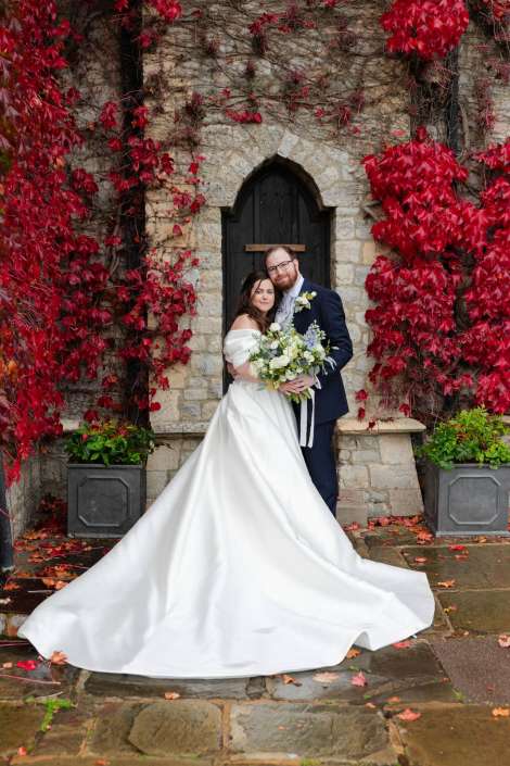 Bride and groom stand together in front of a stone wall with an arched door, surrounded by red ivy, the bride holding a white bouquet.