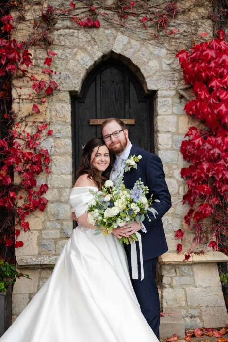 Bride and groom in wedding attire pose together in front of a stone arched door covered with red ivy, the bride holding a white and green bouquet.