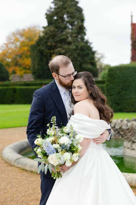 Bride in an off-shoulder white wedding dress and a groom in a navy suit embrace in a garden, as he kisses her on the temple while she holds a bouquet of white and blue flowers.