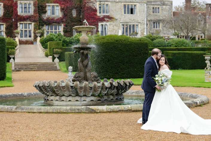 Bride and groom in wedding attire embrace beside a large ornamental fountain in a formal garden with a historic stone mansion in the background.