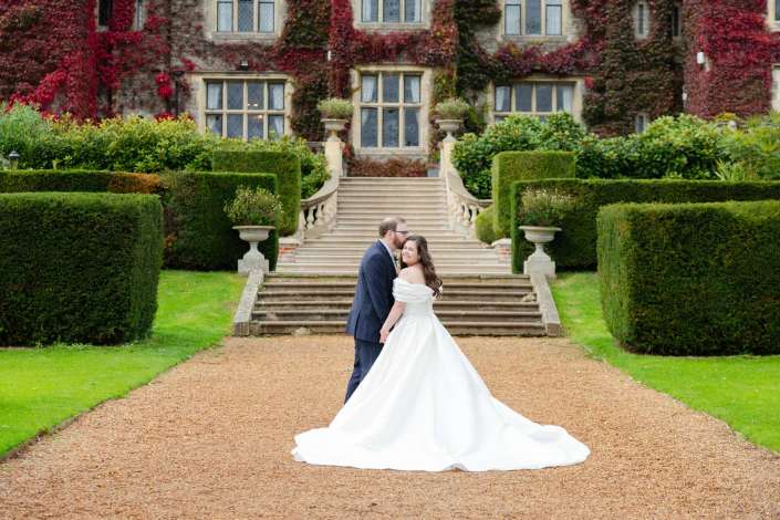 Bride and groom embrace on a gravel path in front of a grand ivy-covered mansion with stone steps and manicured hedges.