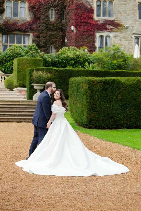 A bride in a long white wedding gown walks hand in hand with a groom in a navy suit along a gravel path in a formal garden with trimmed hedges and a stone building in the background.