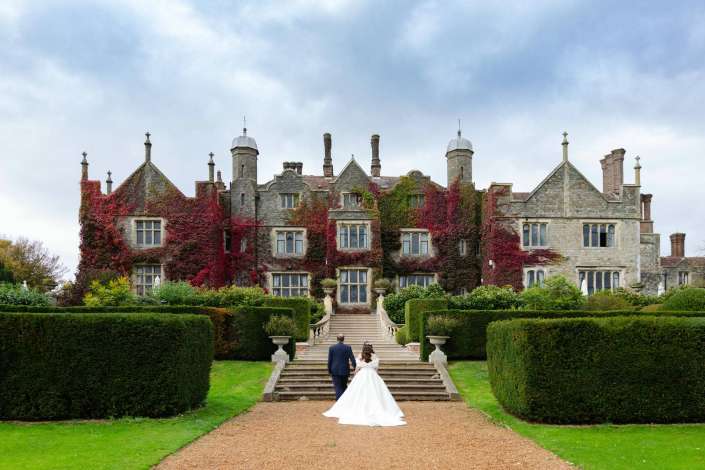 Bride and groom walk up the stone steps toward an ivy-covered historic manor with manicured hedges and a cloudy sky.