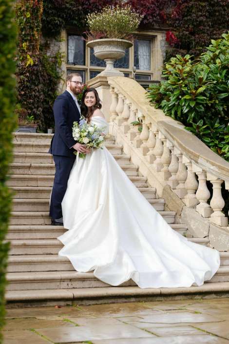 Bride in a white wedding gown and groom in a navy suit stand together on a stone staircase, holding a bouquet amid lush greenery.