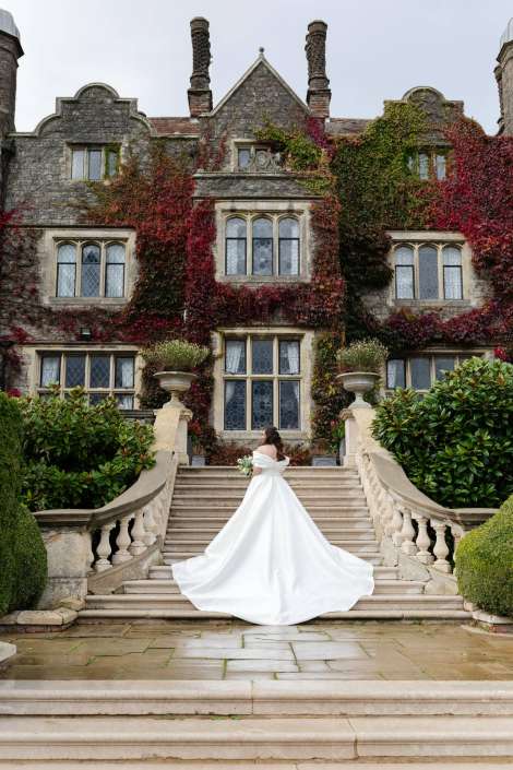 A bride in a flowing white wedding gown holds a bouquet as she stands on a grand stone staircase in front of an ivy-covered historic mansion.