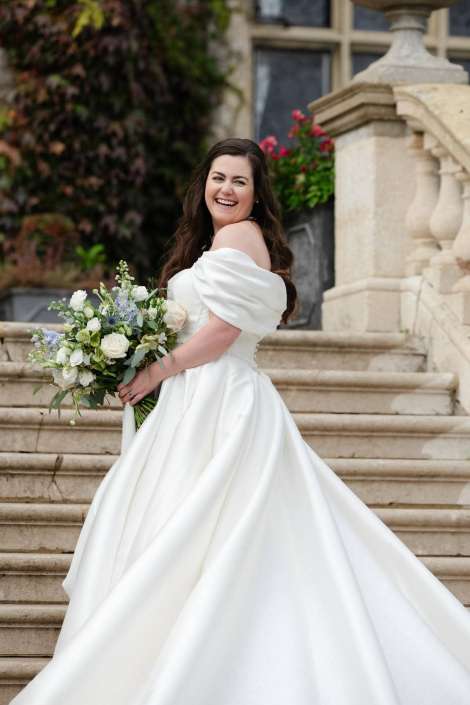 Smiling bride in an off-the-shoulder white wedding dress holding a bouquet on stone steps.