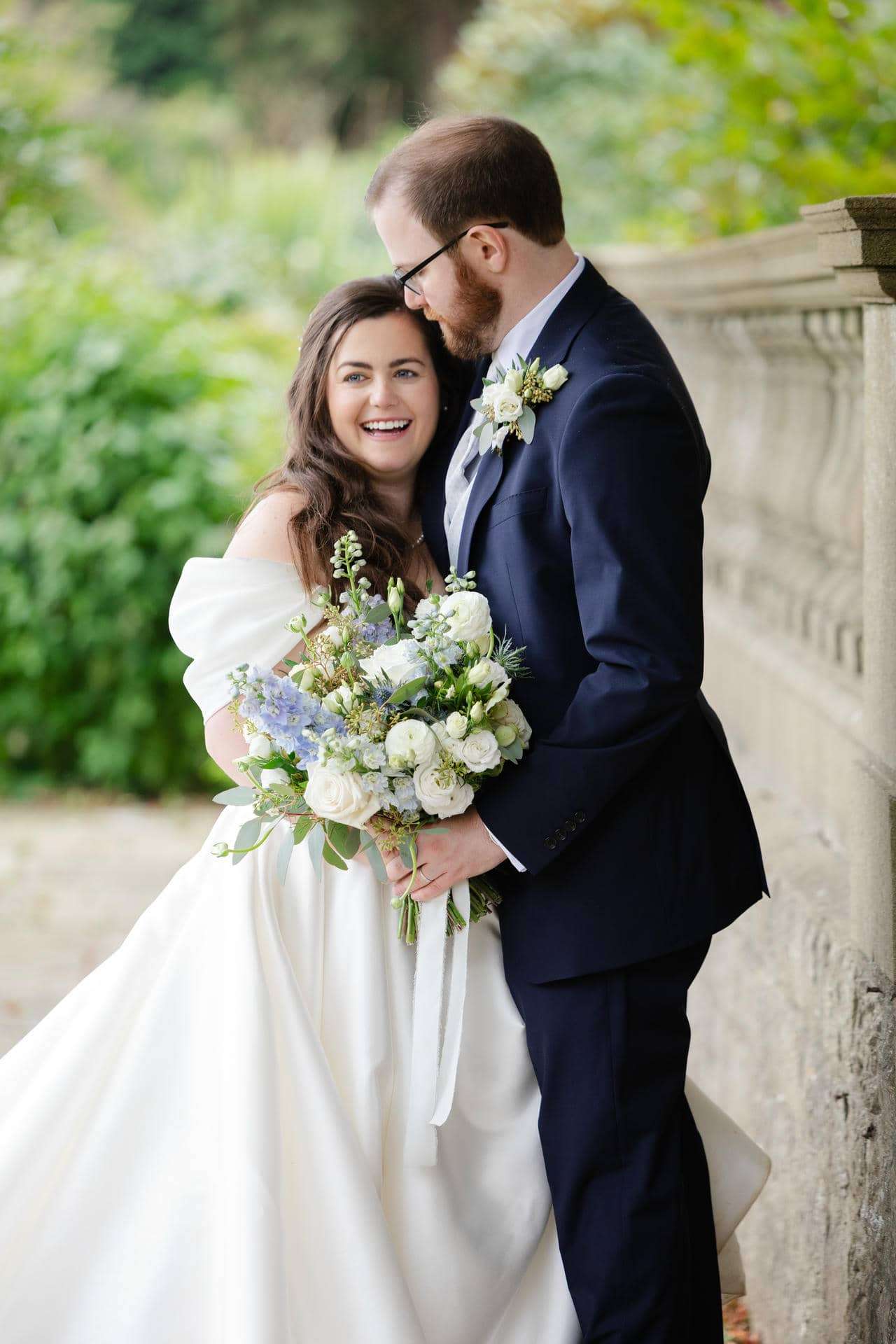 Smiling bride in a white wedding dress holds a bouquet of white and blue flowers beside a groom in a navy suit outdoors.