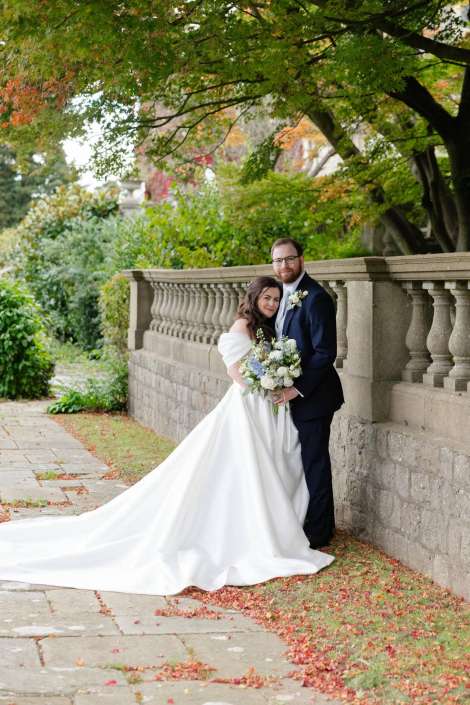 A bride in a white wedding gown with a long train and a bouquet of white flowers stands beside a groom in a navy suit by a stone balustrade in a lush autumn garden.