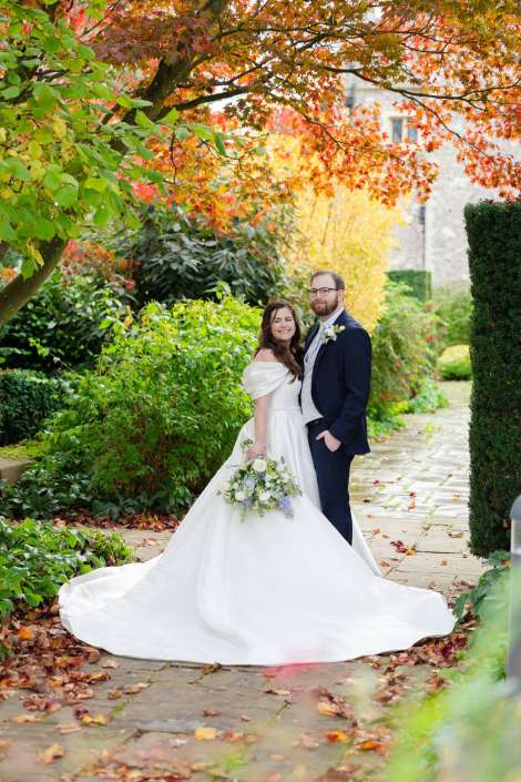 Bride in a white off-the-shoulder gown holds a bouquet beside a groom in a dark suit on a leaf-strewn garden path beneath vibrant autumn foliage.