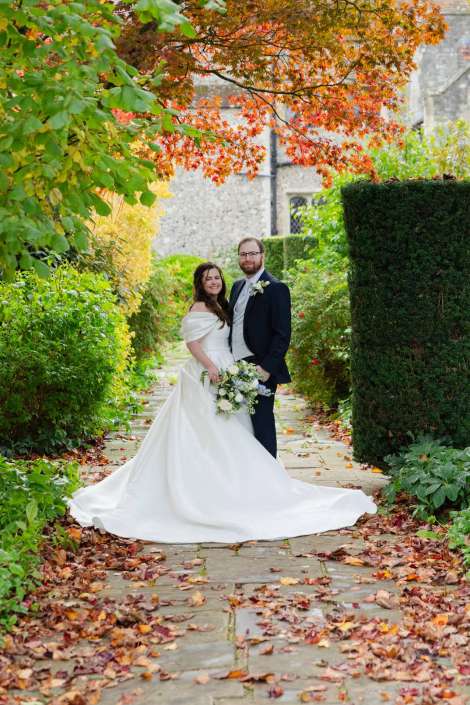 A bride in a white wedding gown and a groom in a dark suit stand together on a stone garden path surrounded by vibrant autumn leaves and greenery.