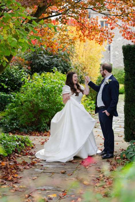 A bride in a white gown and a groom in a navy suit share a joyful moment on a stone path amid vibrant autumn foliage.